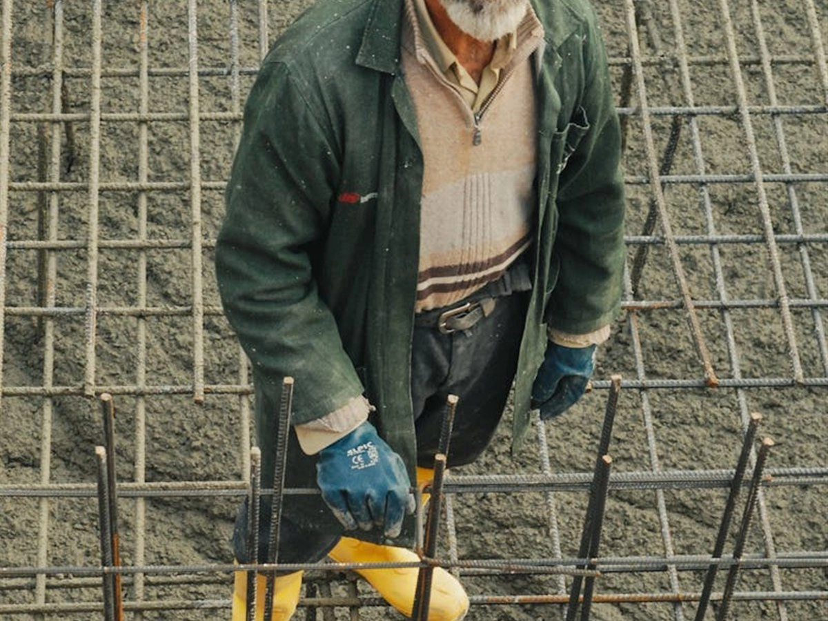 Man in groene jas en gele rubberen laarzen bij wapeningsstaal op een betonvloer